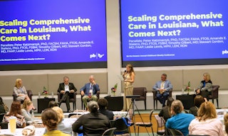 Conference room with tables and attendees seated, listening to an expert panel of speakers on a stage