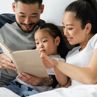 smiling asian parents reading book to toddler daughter in bed